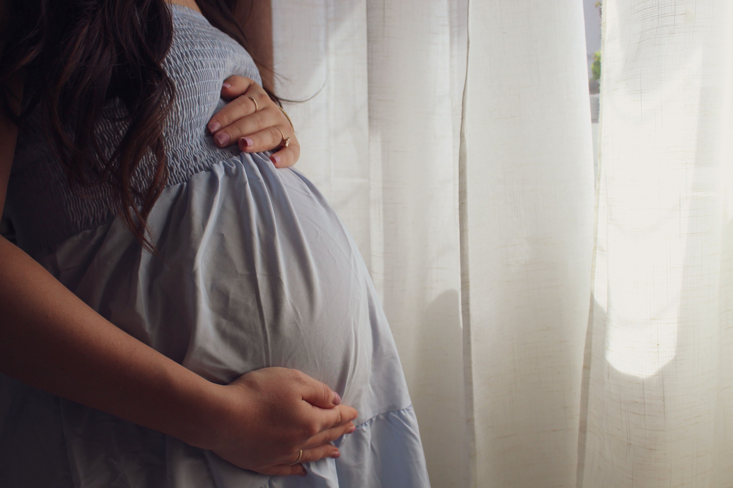 Gravid kvinne i lyseblå kjole holder hendene om magen ved et vindu med lyse gardiner